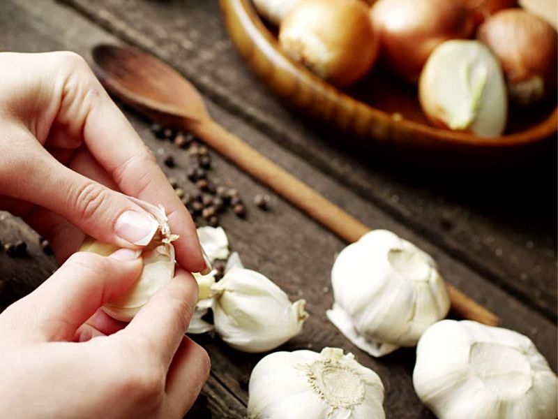 osoba ljušti beli luk, u pozadini činija sa crnim lukom i na stolu par zrnaca bibera